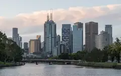 Central Melbourne skyscrapers above the Yarra River, with a bridge and trees on the bank in late afternoon light